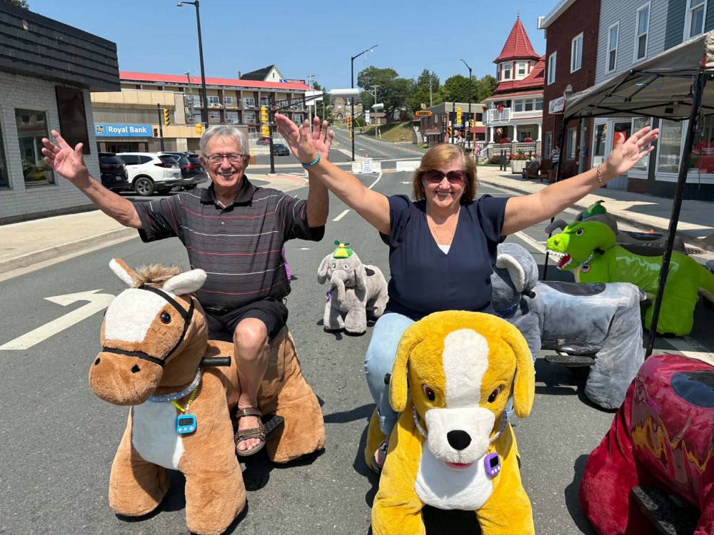 Two adults cheering while riding the Big Horse and Zoomy Doggy at a downtown street event